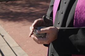 A person wearing a black and purple clergy robe is holding a small glass container with a gray substance, possibly ash. The hands are prominently visible with light highlighting the wrinkles and contours. The background consists of a brick surface with shadows cast, suggesting an outdoor setting.