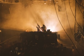 A dimly lit concert venue with smoky ambiance where two DJs are performing on stage. The audience is gathered in front, partly visible in the shadows. The DJs are illuminated by soft stage lights, creating silhouettes against the smoky background. Several electronic equipment pieces, including a laptop, are placed on the table.