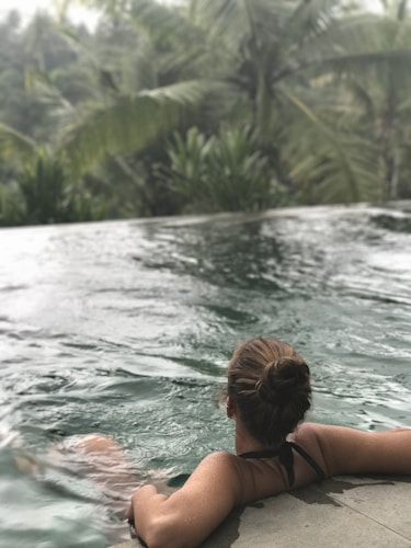 A woman with her hair in a bun is relaxing in an infinity pool overlooking a lush, tropical forest. The water in the pool is still and reflects the surrounding greenery.