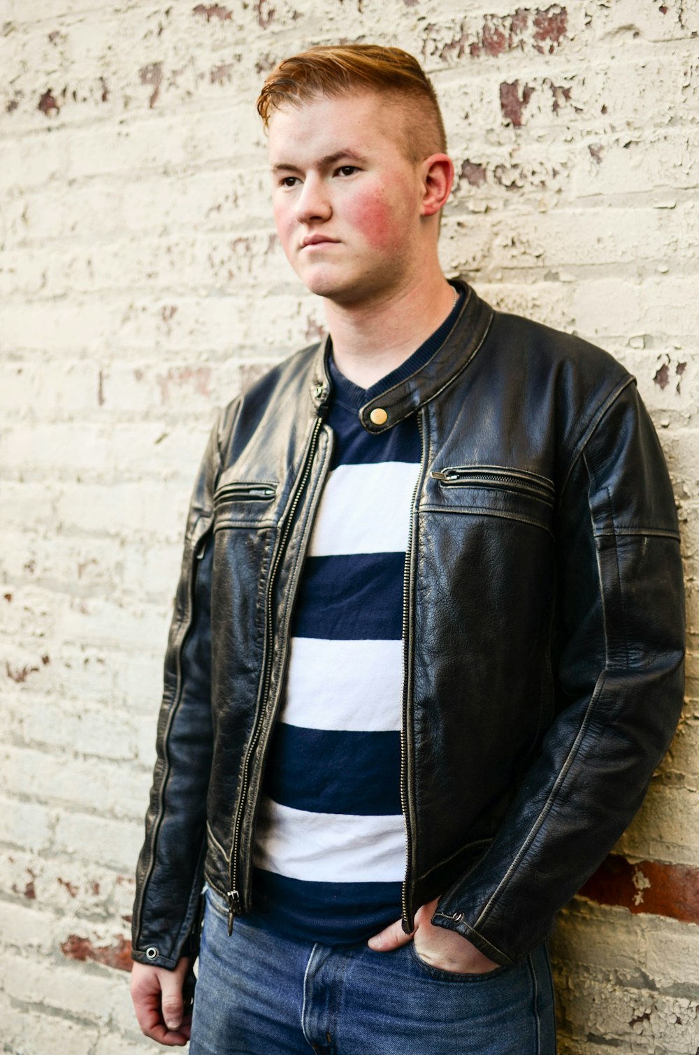 a man in a leather jacket leaning against a brick wall