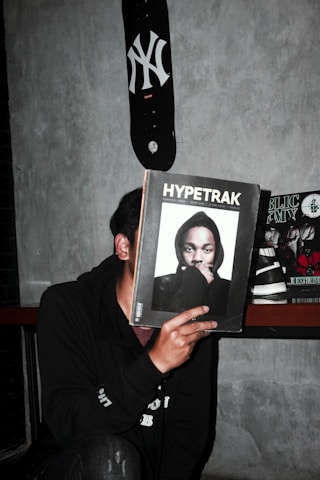 A person is holding a magazine titled 'Hypetrak' in front of their face, obscuring their identity. The magazine cover features a man wearing a hooded sweatshirt. A skateboard with a New York Yankees logo is mounted on the wall above. Other magazines are visible in the background on a shelf. The setting appears to be indoors against a gray concrete wall.
