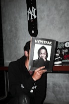 A person is holding a magazine titled 'Hypetrak' in front of their face, obscuring their identity. The magazine cover features a man wearing a hooded sweatshirt. A skateboard with a New York Yankees logo is mounted on the wall above. Other magazines are visible in the background on a shelf. The setting appears to be indoors against a gray concrete wall.