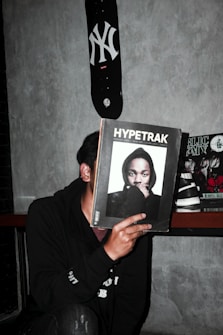 A person is holding a magazine titled 'Hypetrak' in front of their face, obscuring their identity. The magazine cover features a man wearing a hooded sweatshirt. A skateboard with a New York Yankees logo is mounted on the wall above. Other magazines are visible in the background on a shelf. The setting appears to be indoors against a gray concrete wall.