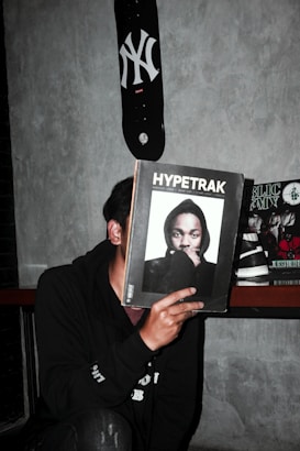 A person is holding a magazine titled 'Hypetrak' in front of their face, obscuring their identity. The magazine cover features a man wearing a hooded sweatshirt. A skateboard with a New York Yankees logo is mounted on the wall above. Other magazines are visible in the background on a shelf. The setting appears to be indoors against a gray concrete wall.