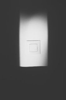 A white light switch is centered on a wall, surrounded by a play of light and shadow. The image is in black and white, creating a minimalist and stark contrast between the illuminated switch and the darker areas.