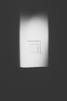 A white light switch is centered on a wall, surrounded by a play of light and shadow. The image is in black and white, creating a minimalist and stark contrast between the illuminated switch and the darker areas.