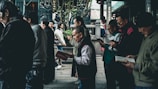 A group of people stands close together, predominantly older adults, each holding and seemingly reading books or pamphlets. The setting appears to be an outdoor area with architectural elements that suggest a traditional or historical location, possibly with cultural or religious significance. The atmosphere is calm, and the individuals focus on their reading material.