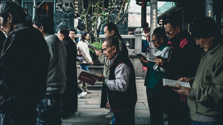 A group of people stands close together, predominantly older adults, each holding and seemingly reading books or pamphlets. The setting appears to be an outdoor area with architectural elements that suggest a traditional or historical location, possibly with cultural or religious significance. The atmosphere is calm, and the individuals focus on their reading material.