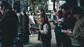 A group of people stands close together, predominantly older adults, each holding and seemingly reading books or pamphlets. The setting appears to be an outdoor area with architectural elements that suggest a traditional or historical location, possibly with cultural or religious significance. The atmosphere is calm, and the individuals focus on their reading material.