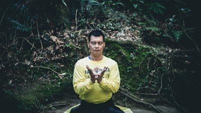 A man is sitting cross-legged in a meditative pose, wearing a yellow shirt with blue writing. He has his eyes closed, and his hands are positioned in a mudra. The background consists of a lush, green wooded area with moss and fallen leaves.