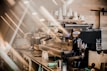 A professional espresso machine stands on a countertop surrounded by coffee-making equipment in a cozy, softly-lit café. Light beams create a warm, inviting atmosphere, highlighting the metallic textures of the machine and nearby glassware.