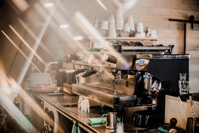 A professional espresso machine stands on a countertop surrounded by coffee-making equipment in a cozy, softly-lit café. Light beams create a warm, inviting atmosphere, highlighting the metallic textures of the machine and nearby glassware.