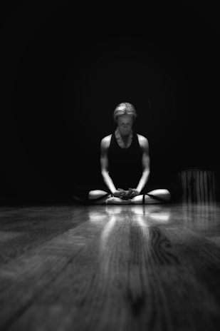 A person is sitting cross-legged on a wooden floor with a focused expression. The setting is dark with a spotlight creating reflections on the polished surface. The scene suggests solitude and introspection.