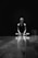 A person is sitting cross-legged on a wooden floor with a focused expression. The setting is dark with a spotlight creating reflections on the polished surface. The scene suggests solitude and introspection.