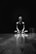A person is sitting cross-legged on a wooden floor with a focused expression. The setting is dark with a spotlight creating reflections on the polished surface. The scene suggests solitude and introspection.