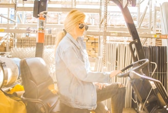 A person wearing sunglasses and a denim jacket is seated on a forklift in a warehouse setting. The environment is industrial, with metal shelves and various materials like metal pipes and bricks in the background. Sunlight is streaming in, creating a bright and warm ambiance.