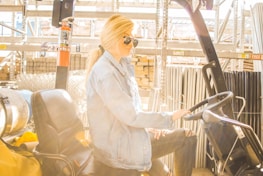 A person wearing sunglasses and a denim jacket is seated on a forklift in a warehouse setting. The environment is industrial, with metal shelves and various materials like metal pipes and bricks in the background. Sunlight is streaming in, creating a bright and warm ambiance.
