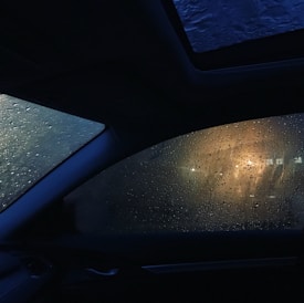 Water droplets cover a car window, refracting light from a warm, glowing source outside. The interior of the vehicle is dimly lit, with the dark silhouette of the car's frame visible against the moist, glistening glass.