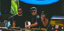Three individuals are gathered behind a DJ booth, with two men and one woman. They are dressed in casual attire and sunglasses, indicating a sunny outdoor setting. The man in the center is operating the DJ equipment, while the other two appear to be enjoying the music. There are palm leaves visible in the background, suggesting a tropical location.