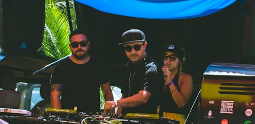Three individuals are gathered behind a DJ booth, with two men and one woman. They are dressed in casual attire and sunglasses, indicating a sunny outdoor setting. The man in the center is operating the DJ equipment, while the other two appear to be enjoying the music. There are palm leaves visible in the background, suggesting a tropical location.