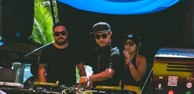 Three individuals are gathered behind a DJ booth, with two men and one woman. They are dressed in casual attire and sunglasses, indicating a sunny outdoor setting. The man in the center is operating the DJ equipment, while the other two appear to be enjoying the music. There are palm leaves visible in the background, suggesting a tropical location.