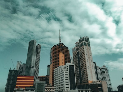 A diverse skyline featuring several tall buildings against a cloudy sky, with a mix of modern and older architectural styles. The structures vary in height and design, with one notable building having an orange facade.