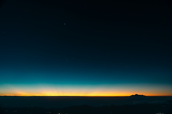 A striking sunset or sunrise over a mountainous horizon with a wide expanse of sky transitioning from deep blue to a warm orange near the horizon. The silhouette of a mountain range is visible against the bright glow of the sky.