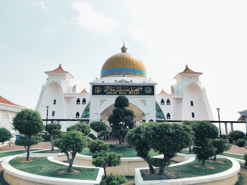 A grand mosque with a prominent gold dome, surrounded by carefully manicured trees and shrubs. The building features white walls with intricate architectural elements and ornate windows. There is a sign with Arabic script at the entrance.