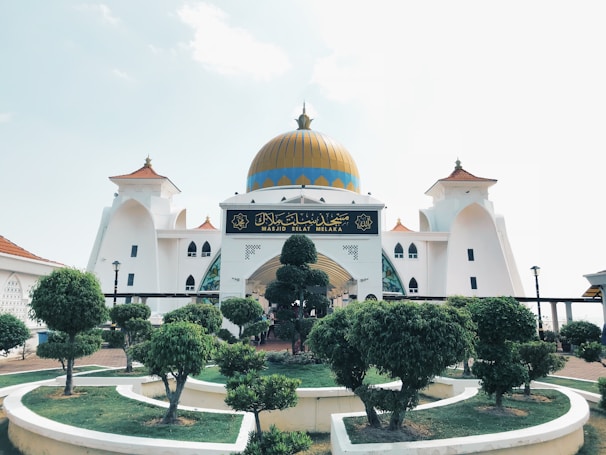 A grand mosque with a prominent gold dome, surrounded by carefully manicured trees and shrubs. The building features white walls with intricate architectural elements and ornate windows. There is a sign with Arabic script at the entrance.