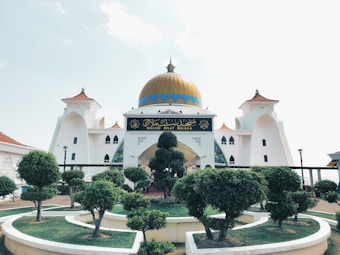 A grand mosque with a prominent gold dome, surrounded by carefully manicured trees and shrubs. The building features white walls with intricate architectural elements and ornate windows. There is a sign with Arabic script at the entrance.