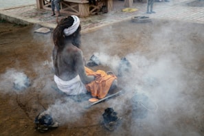 A person with long hair sits in a meditative pose on the ground, surrounded by smoking embers. The individual is wrapped in white fabric and holds an orange cloth. Nearby, there are other objects including metal containers. The setting appears to be outdoors, with a sandy ground and stone pathways visible in the background.