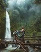 photo of man and woman holding tires and walking on bridge near waterfalls