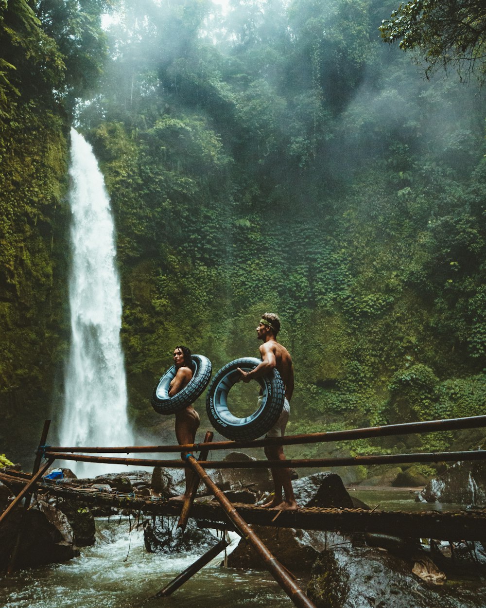 photo of man and woman holding tires and walking on bridge near waterfalls