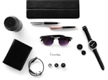 Various everyday items neatly arranged on a white surface, including a black wallet, wristwatch, sunglasses, pen, notebook, portable speaker, bracelet, ink bottle caps, and a card labeled 'Essentials.'