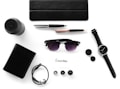 Various everyday items neatly arranged on a white surface, including a black wallet, wristwatch, sunglasses, pen, notebook, portable speaker, bracelet, ink bottle caps, and a card labeled 'Essentials.'