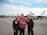Three men are standing on an airfield with a line of fighter jets behind them. Two are dressed in black pilot uniforms with a patch and insignia, while the man in the middle is wearing a red shirt and glasses. They appear to be smiling and shaking hands.