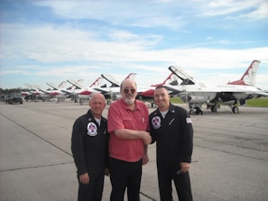 Three men are standing on an airfield with a line of fighter jets behind them. Two are dressed in black pilot uniforms with a patch and insignia, while the man in the middle is wearing a red shirt and glasses. They appear to be smiling and shaking hands.