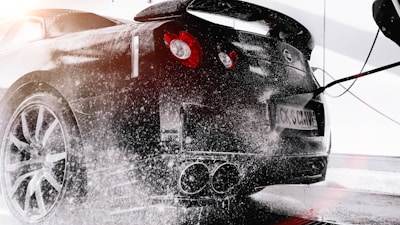 A black sports car is undergoing a wash, with soap suds covering its surface. The rear of the vehicle is visible, including red taillights and dual exhaust pipes. Water is being sprayed on the car from a hose, creating splashes and reflections on the glossy exterior. The scene is brightly lit, suggesting a clean and fresh environment.