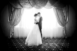 A romantic black and white scene featuring a bride and groom sharing an intimate moment. They are standing close together, facing each other, in front of a large, elegantly draped window. The backlighting creates a silhouette effect, highlighting the grace of the bridal gown and the couple's embrace.