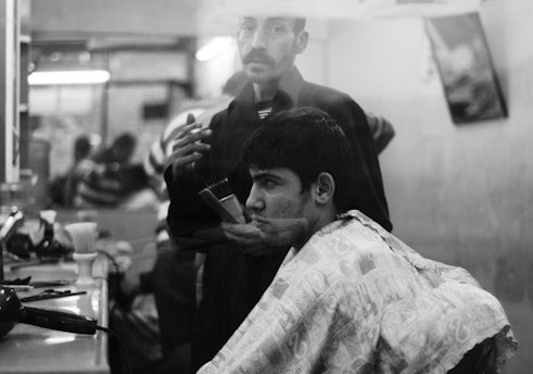 A black and white image of a man getting a haircut in a barber shop. The barber, holding a comb and scissors, stands behind him, focusing on the task. The background shows reflections and blurred figures, suggesting a busy environment.