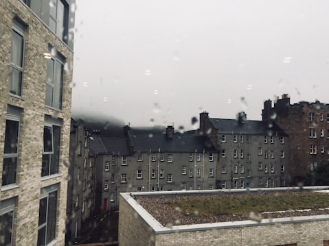 A view through a rain-soaked window of several multi-story buildings with slate roofs. The architecture is traditional with brickwork and multiple windows. The sky is overcast, and there is a sense of mist or fog in the distance, giving the scene a gloomy and moody atmosphere.