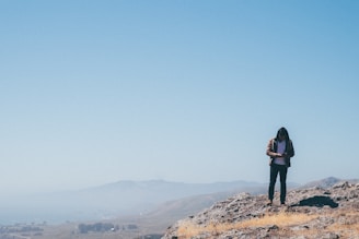 A person stands on a rocky hill, looking down at a phone or a small device in their hands. The background features a vast, slightly hazy landscape of rolling hills under a clear blue sky. The scene suggests solitude and contemplation in nature.