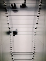 A glass staircase viewed from below, with silhouettes of people walking overhead. The design features evenly spaced steps supported by small fixtures on each side, casting bold shadows on the surface.