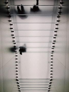 A glass staircase viewed from below, with silhouettes of people walking overhead. The design features evenly spaced steps supported by small fixtures on each side, casting bold shadows on the surface.