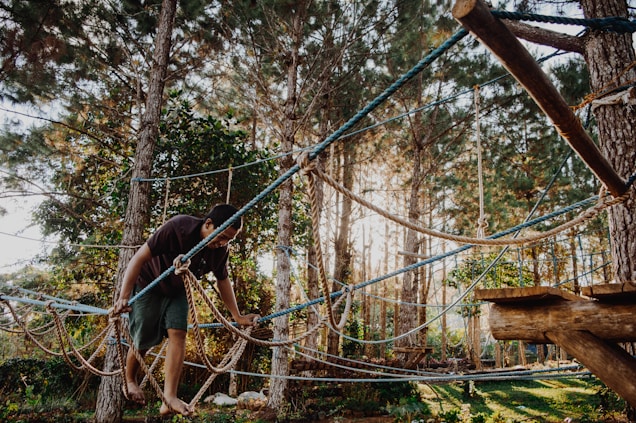 A person is navigating across a high rope course in the middle of a forest. The forest consists of tall trees with green foliage. The person is barefoot and using both hands to hold onto the ropes for balance. The atmosphere is bright with sunlight filtering through the trees.