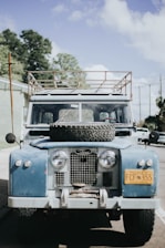 An old blue Land Rover is parked on a street, with a spare tire mounted on its hood. The vehicle has a roof rack and a distinct yellow license plate from Alaska. The scene is set against a backdrop of trees and a partly cloudy sky.