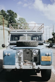 An old blue Land Rover is parked on a street, with a spare tire mounted on its hood. The vehicle has a roof rack and a distinct yellow license plate from Alaska. The scene is set against a backdrop of trees and a partly cloudy sky.