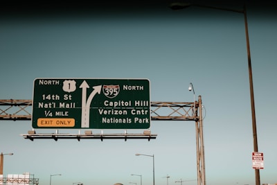 A highway sign directs drivers toward the National Mall and Capitol Hill, with arrows indicating separate routes. The sign is mounted on a metal framework, with a clear blue sky in the background. Additional street lamps and a smaller sign about snow emergency routes are visible.