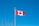 A Canadian flag with a red maple leaf on a white background, bordered by red vertical bands, is flying on a flagpole against a clear blue sky. Bare tree branches are visible at the bottom left corner.
