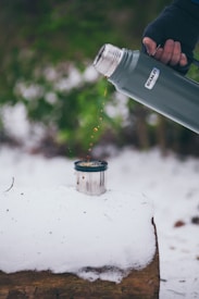 A person wearing a glove is pouring liquid from a green thermos into a small metal cup. The cup is placed on top of a snow-covered surface in an outdoor setting. The background is blurred with hints of greenery.